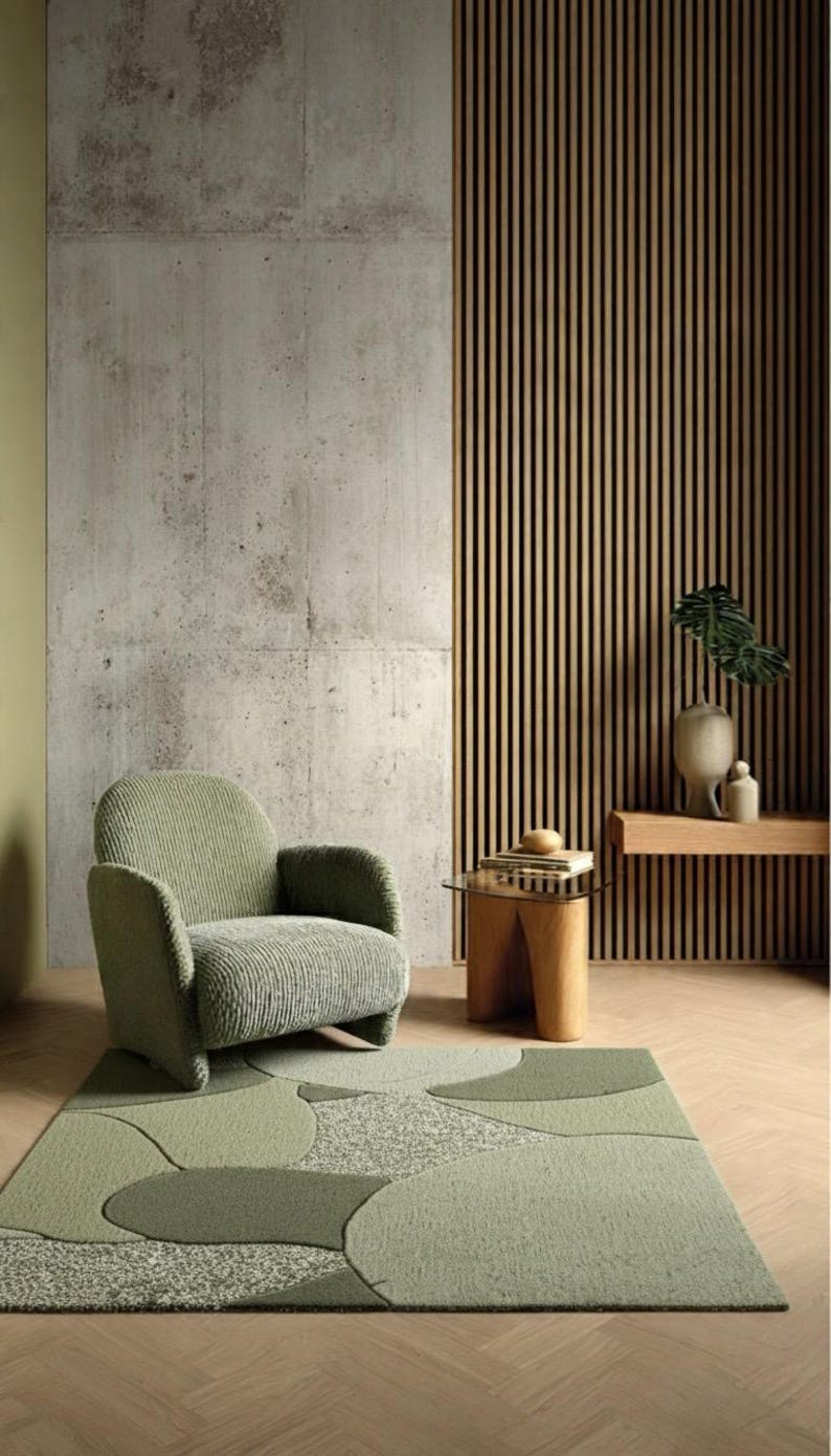 May include: A sage green corduroy armchair with rounded arms and a backrest, positioned on a rectangular area rug featuring a green abstract pattern of overlapping circles and textured sections, set against a concrete and wood slat wall.