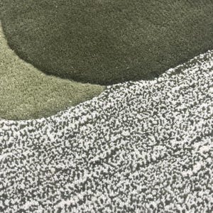 May include: Close-up of a textured rug with a layered design. The rug features shades of green and white, with a soft, plush appearance. The design includes a large, circular shape in a darker green.