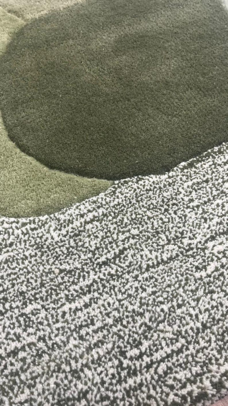 May include: Close-up of a textured rug with a layered design. The rug features shades of green and white, with a soft, plush appearance. The design includes a large, circular shape in a darker green.