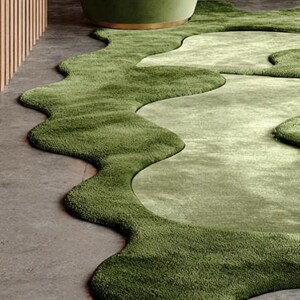 May include: A green, organic-shaped rug with varying shades of green, resembling a landscape. The rug is placed on a concrete floor, with a green armchair in the background. The design creates a flowing, natural aesthetic.