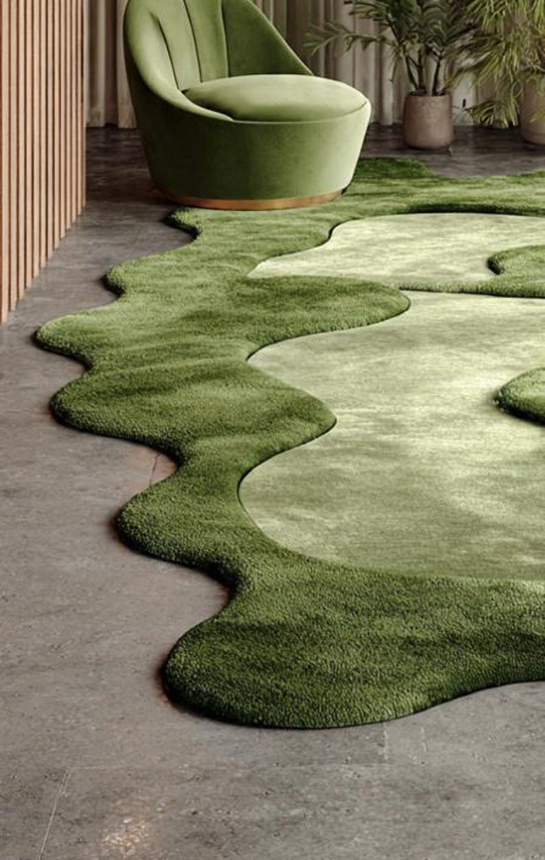 May include: A green, organic-shaped rug with varying shades of green, resembling a landscape. The rug is placed on a concrete floor, with a green armchair in the background. The design creates a flowing, natural aesthetic.