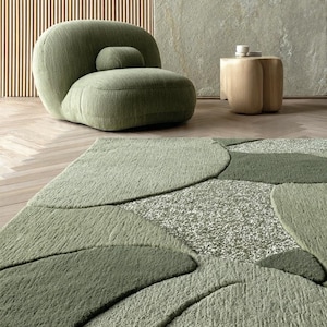 Puede incluir: Una escena de salón moderno con un sillón grande, verde oliva, redondeado y un pequeño cojín a juego. Una mesa auxiliar de madera clara y una alfombra estampada en tonos verdes completan el espacio. La alfombra tiene un diseño orgánico único.
