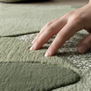 May include: Close-up of a green rug with a textured, organic design. The rug features varying shades of green, creating a layered effect. The surface appears soft and plush, with a hand gently touching the rug's surface.