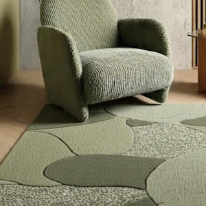 May include: A sage green armchair with a textured, ribbed fabric sits on a patterned rug. The rug features a design of overlapping shapes in various shades of green. The room has a neutral color palette.