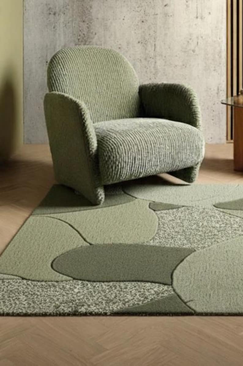 May include: A sage green armchair with a textured, ribbed fabric sits on a patterned rug. The rug features a design of overlapping shapes in various shades of green. The room has a neutral color palette.