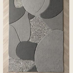 May include: A rectangular area rug with a modern design. The rug features a pattern of overlapping shapes in various shades of gray, with a speckled, lighter gray section in the center. The word "Grey" is printed at the bottom.