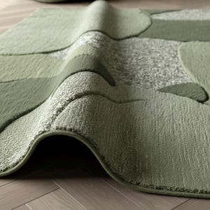 May include: A sage green area rug featuring a textured, abstract pattern of rounded shapes and speckled sections. The rug is folded over, revealing a thick, plush pile construction with varying surface heights and a soft, tufted finish.