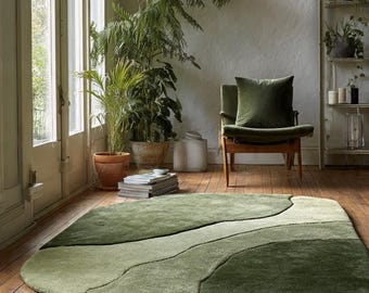 May include: A green area rug with an abstract design, featuring varying shades of green. The rug has a soft, plush texture and is placed on a wooden floor. The room also includes a green chair, plants, and a window.