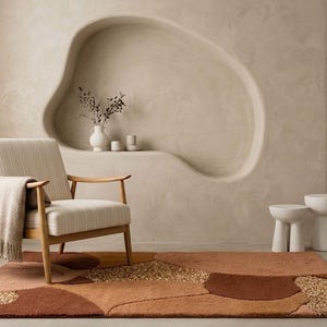 May include: A modern living room scene featuring a beige armchair with a striped seat and wooden frame, a patterned rug in shades of brown and beige, and a decorative wall niche. Two small white stools and potted plants complete the decor.