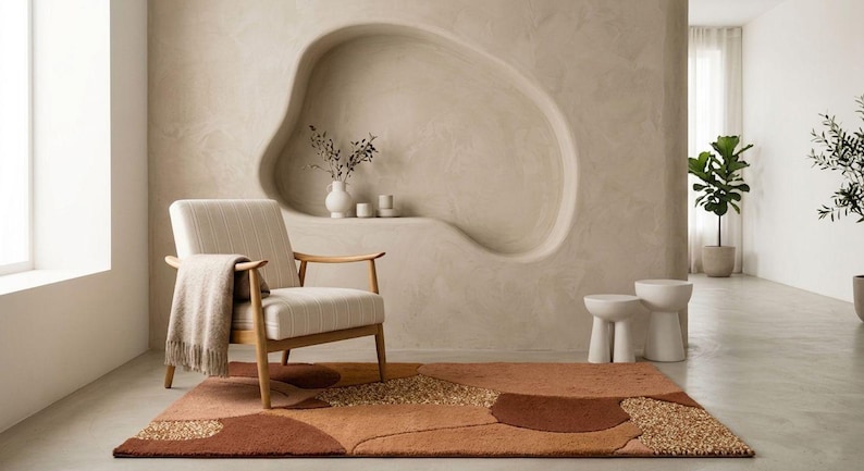 May include: A modern living room scene featuring a beige armchair with a striped seat and wooden frame, a patterned rug in shades of brown and beige, and a decorative wall niche. Two small white stools and potted plants complete the decor.