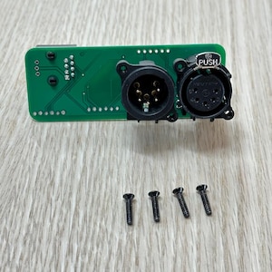 May include: A green circuit board with two black circular connectors and four black screws. One connector has the word "PUSH" printed on it. The connectors have multiple pins.