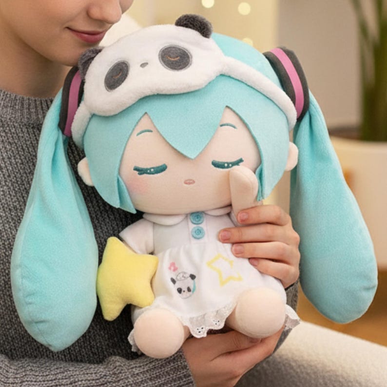 May include: A plush doll with light blue hair and a panda eye mask. The doll wears a white dress with a panda and star design and holds a yellow star. The doll is being held by a person.
