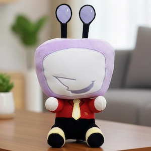 Könnte beinhalten: Ein Plüschtier in Form eines Cartoon-Fernsehers. Das Spielzeug hat einen lila Kopf mit zwei antennenartigen Merkmalen, einen weißen Bildschirm und einen lächelnden Mund. Es trägt ein rotes Hemd, eine schwarze Hose und eine gelbe Krawatte. Das Spielzeug sitzt auf einer Holzoberfläche.