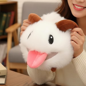Könnte beinhalten: Ein weißes, flauschiges Plüschtier mit braunen Hörnern und einer rosa Zunge. Das Spielzeug hat ein lächelndes Gesicht mit einem schwarzen Auge. Die Person trägt einen cremefarbenen Pullover. Das Spielzeug ist eine Zeichentrickfigur.