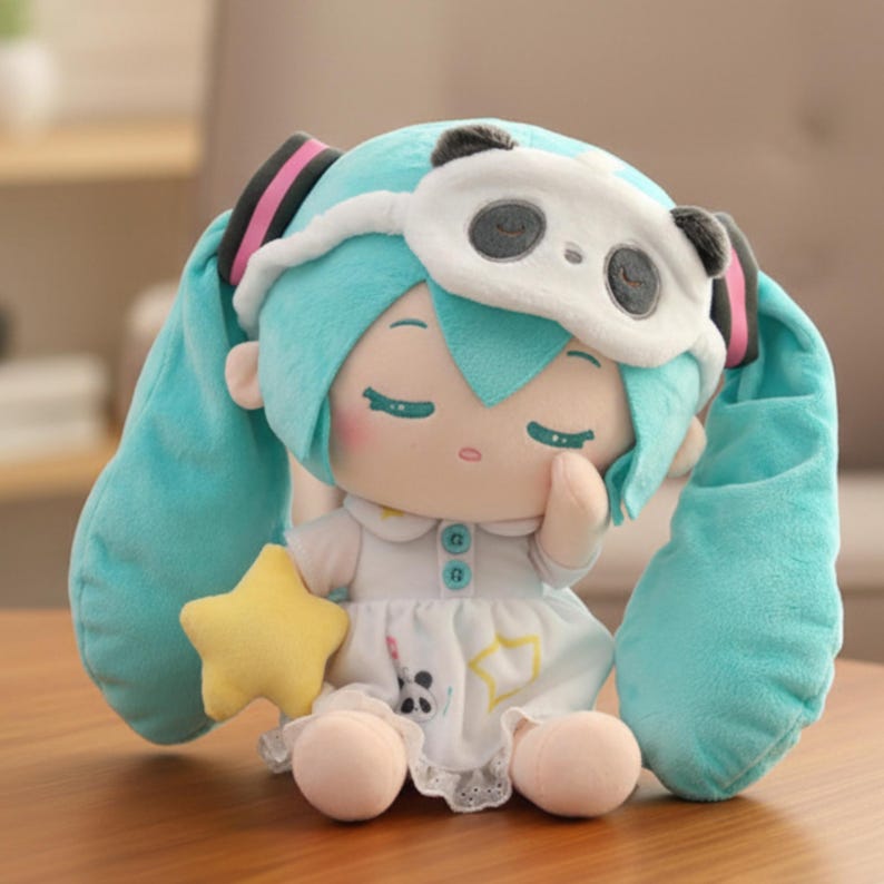 May include: A plush doll with turquoise hair and a panda eye mask. The doll is wearing a white dress with a star and panda print and holding a yellow star. The doll has closed eyes and a peaceful expression.