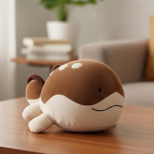 Puede incluir: Un peluche de ballena marrón y blanca. La ballena tiene un cuerpo redondeado con manchas blancas, una boca sonriente y aletas pequeñas. Está sobre una mesa de madera.