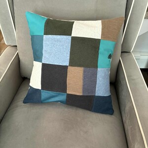 May include: A cushion with a patchwork design, featuring squares of blue, teal, brown, and white fabrics. The cushion is on a grey armchair. The Adidas logo is visible on one of the teal squares.