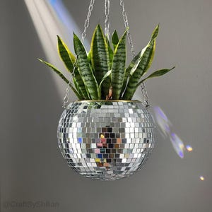May include: A hanging disco ball planter with a snake plant. The spherical planter is covered in small, reflective mirrored tiles. The plant has long, green and yellow striped leaves. The planter hangs from a silver chain against a gray background.