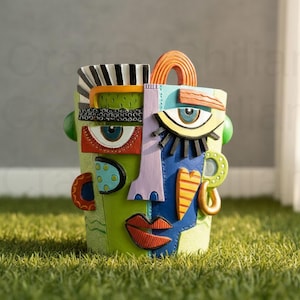 Puede incluir: Una maceta colorida con forma de rostro abstracto con una variedad de formas y patrones geométricos. La maceta presenta una base verde con detalles azules, naranjas y morados, y un gran diseño de ojo. La maceta está sobre hierba verde.