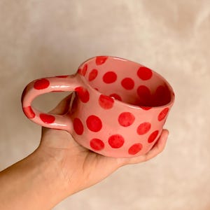 Peut inclure: Une tasse en céramique rose avec des pois rouges, tenue dans une main. La tasse a une grande anse et un intérieur rose. La tasse faite à la main est une pièce unique, idéale pour le thé ou le café. Le design de la tasse est simple mais accrocheur.