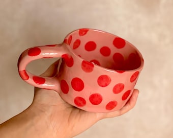 May include: A pink ceramic mug with a large handle and red polka dots. The mug is held in a hand, showcasing its handmade quality. The mug's interior is also pink, creating a cohesive design. This unique mug is perfect for coffee or tea.