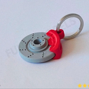 May include: A gray and red brake rotor keychain with a silver key ring. The brake rotor has drilled holes and is paired with a red brake caliper. The keychain is a miniature replica of an automotive brake system.