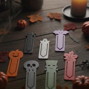 May include: A selection of Halloween-themed bookmarks in various colours and designs, including a pumpkin, ghost, skull, and Frankenstein's monster. The bookmarks are on a wooden surface with leaves, pumpkins, and a lit candle.