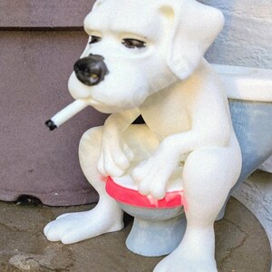 May include: A white figurine of a dog sitting on a toilet, smoking a cigarette. The dog has a black nose and is sitting on a toilet with a red seat. The figurine is on a round, dark surface.