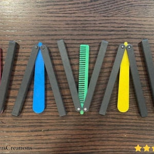 Puede incluir: Una colección de peines mariposa coloridos, cada uno con un peine y un mango de diferente color. Los mangos son de color gris oscuro y los peines vienen en rosa, azul, verde, amarillo y naranja. Los peines se exhiben sobre una superficie de madera.