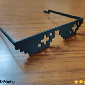 May include: Black pixelated sunglasses with a distinctive design. The glasses feature a unique, blocky aesthetic, with cross-shaped and square cutouts. The arms extend to the sides, and the glasses are set against a wooden surface.