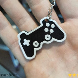 May include: A black and white keychain featuring a video game controller design. The controller has a white outline and details, including buttons and a directional pad. The keychain is attached to a silver chain.