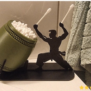 Può includere: Un porta cotton fioc a forma di tamburo verde con la silhouette nera di un artista marziale che tiene i cotton fioc come bacchette. Il supporto è pieno di cotton fioc bianchi.