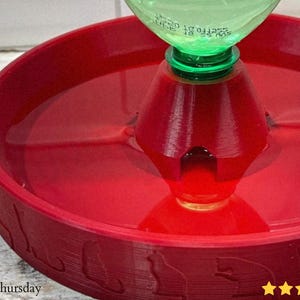 Puede incluir: Una fuente de agua roja para gatos con una tapa de botella de pl&aacute;stico verde. La fuente tiene una base circular con siluetas de gatos alrededor del borde. El agua es visible dentro.
