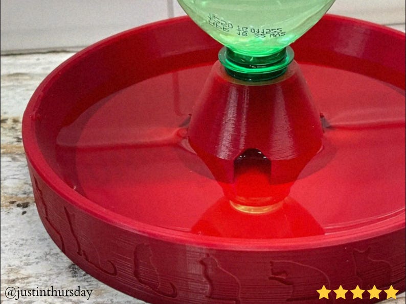 Puede incluir: Una fuente de agua roja para gatos con una tapa de botella de pl&aacute;stico verde. La fuente tiene una base circular con siluetas de gatos alrededor del borde. El agua es visible dentro.