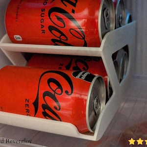May include: A white, two-tiered can organiser inside a fridge, holding several red Coca-Cola Zero cans. The cans are stacked, with the Coca-Cola logo prominently displayed. The organiser is designed to fit inside a fridge.