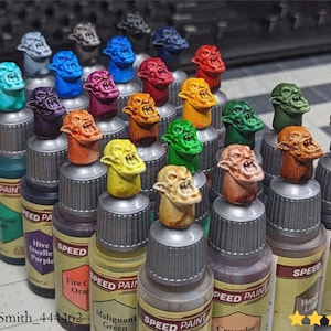 以下が含まれることがあります： さまざまな色のミニチュアモンスターの頭が上に乗った、Speed Paintのボトルのコレクション。ボトルには「Holy White」や「Malignant Green」などのペイント名がラベル表示されています。画像には「@JosiahSmith_444462」というテキストが含まれています。