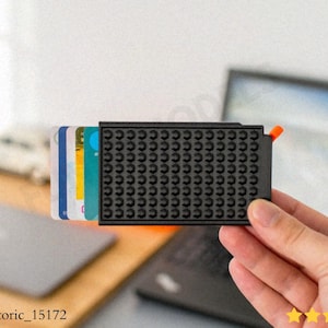 Peut inclure: Un porte-cartes noir avec une surface texturée, contenant plusieurs cartes colorées. Le support a un accent orange et est tenu dans une main. L'arrière-plan comprend un ordinateur portable et une surface en bois.