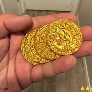 May include: A handful of gold-colored coins with a skull design in the center. The coins have an intricate circular pattern around the skull. The coins are held in a hand.