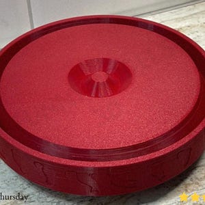 Puede incluir: Un objeto redondo rojo con una superficie texturizada y una zona central elevada. El objeto tiene un borde ancho y plano con un patr&oacute;n decorativo. El dise&ntilde;o es circular y sim&eacute;trico.