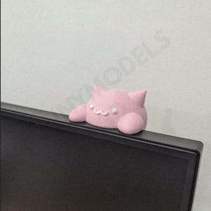 May include: A light pink cat-shaped webcam cover with a smiling face and small ears, placed on a black computer monitor. The background is a plain white wall. A cute and functional accessory.