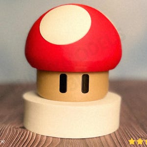May include: A red and white mushroom figurine, reminiscent of a Super Mario Bros. power-up. The mushroom has a red cap with a white circle and a tan base with two black rectangular eye-like shapes. It sits on a white cylindrical base.