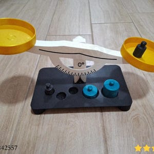 May include: A wooden balance scale with yellow pans and a black base. The scale has a white arm and a black base with various weights, including blue cylinders and black cylinders. The scale is designed for educational purposes.