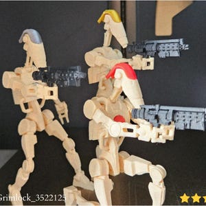 May include: Three beige-colored action figures of battle droids, each holding a gray blaster rifle. The figures have articulated joints and are posed in dynamic stances. The figures have red and yellow accents. The image includes the text "@KingGrimlock_3522125".