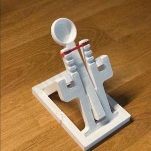 May include: A white 3D-printed trebuchet model with a red rubber band. The model has a rectangular base and a pivoting arm with a small cup. The text "@highpoly" is visible.