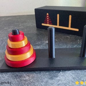 Puede incluir: Un juego de rompecabezas de madera con anillos rojos y amarillos, una base negra con dos postes y una caja negra con un diseño similar. El juego está diseñado para el juego educativo.