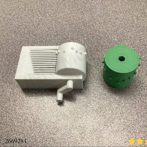 May include: A 3D-printed, off-white, rectangular object with a cylindrical component and a handle, next to a green cylindrical object. The rectangular object has horizontal lines and a cylindrical top with small protrusions. The green cylinder has a central hole.