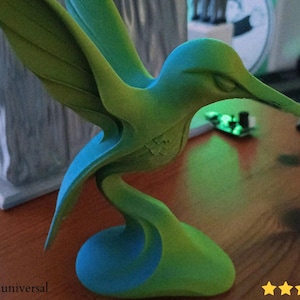 May include: A sculpted hummingbird figurine in shades of green and blue. The bird is posed mid-flight, with its wings extended. The sculpture has a smooth, flowing design and is set on a wooden surface.