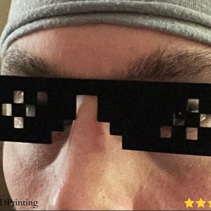 May include: Black pixelated sunglasses with a mosaic design. The sunglasses are worn by a person wearing a gray beanie. The image includes the text "@Keks3DPrinting" and five gold stars.