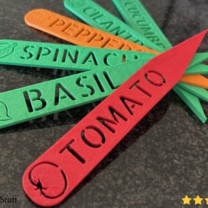 May include: A collection of colorful garden plant markers. The markers are red, green, and orange, with cut-out lettering indicating the plant type. The red marker reads "TOMATO" and has a tomato illustration. Other markers include "BASIL", "SPINACH", "PEPPER", and "CUCUMBER".