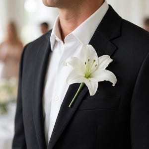 May include: A black suit jacket worn over a crisp white collared shirt, featuring a single white lily flower pinned to the lapel as a boutonniere. The flower has prominent green stamens and is positioned on the left side of the chest.